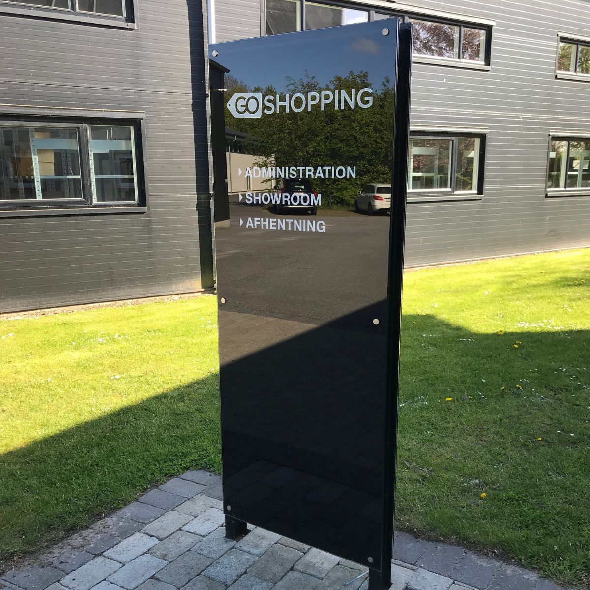 Fritstående sort informationsskilt med oversigt over shopping, administration, showroom og parkering, placeret udendørs foran kontorbygning