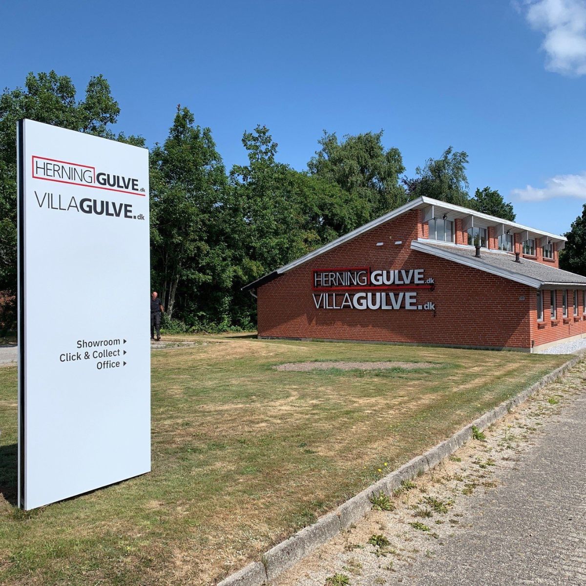 Udendørs wayfinding- og områdeskilt med teksten ‘Herning Gulve, VillaGulve’, placeret ved indkørsel foran erhvervsbygning