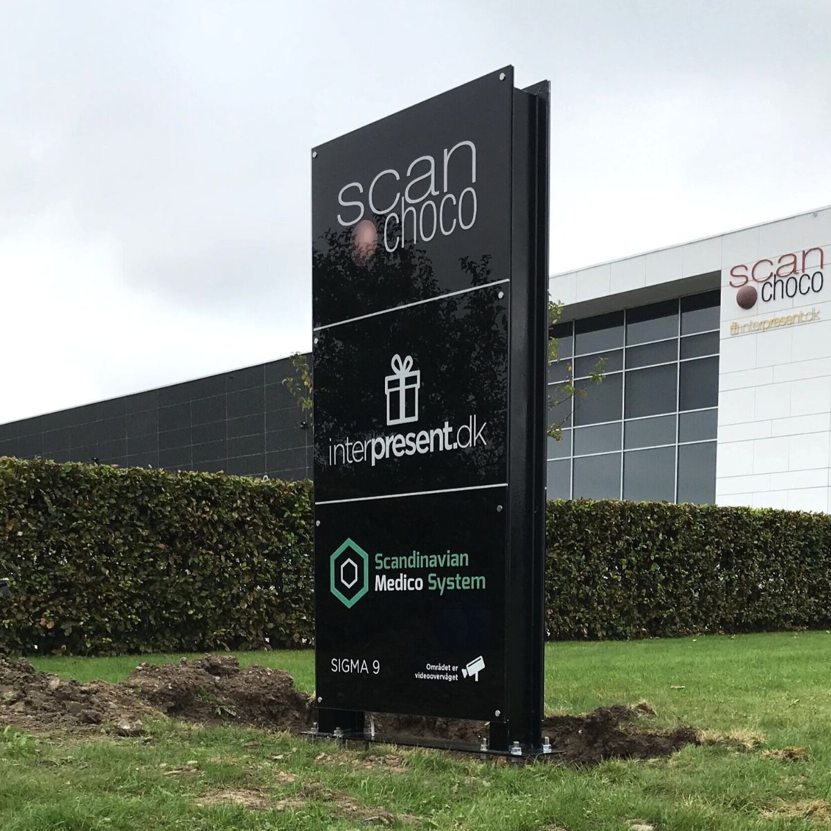 Udendørs pylon- og wayfinding-skilt med virksomhedslogoer og oversigt over erhvervslejemål, placeret foran erhvervsbygning