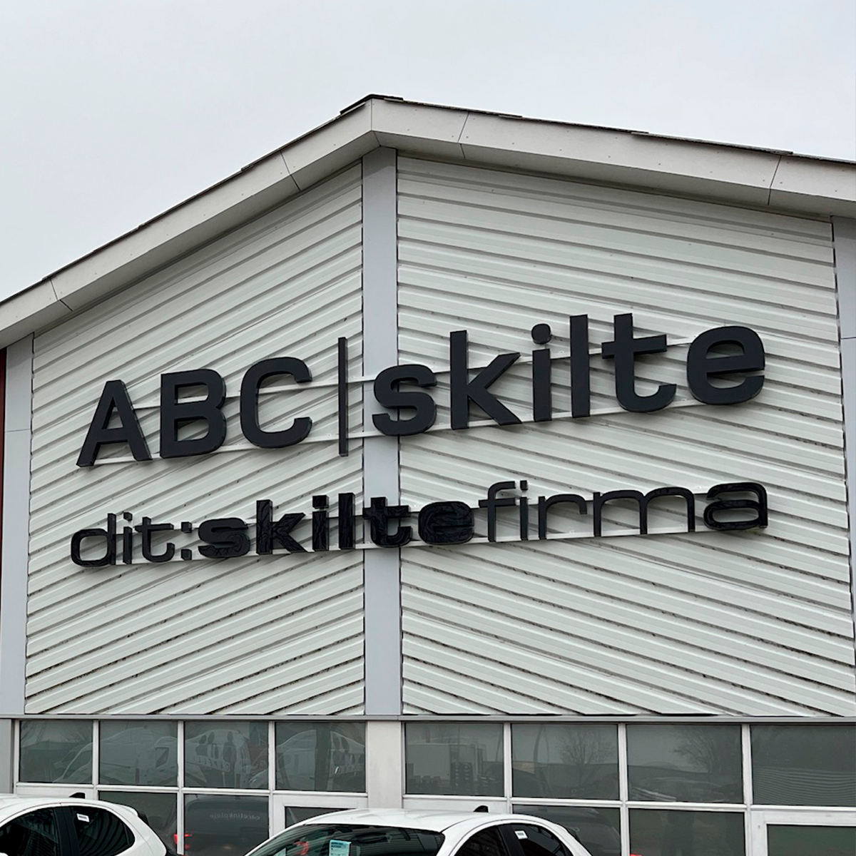 Bygning i hvid træ med facadeskilte i sort "ABC Skilte" og "dit:skiltefirma" 