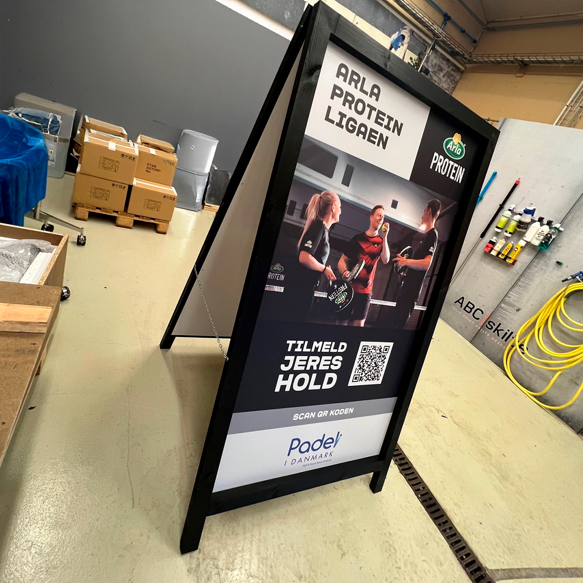 Sort fritstående reklameskilt (A-skilt) med Padel-reklame, placeret i et lager- eller værkstedsmiljø med kasser og udstyr i baggrunden