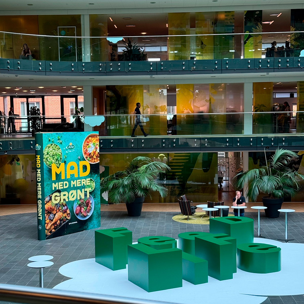 Indendørs butiks- eller eventinstallation med grønne siddeelementer og grafisk skilt i et moderne atriummiljø