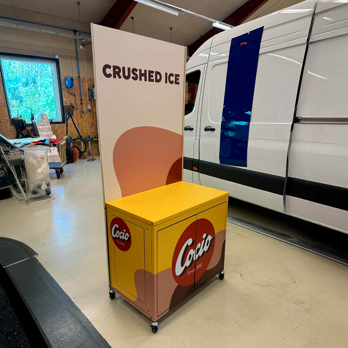 Gul Coca-Cola salgsdisplay med teksten ‘CRUSHED ICE’, placeret i et lager- eller produktionsmiljø ved siden af en hvid varebil