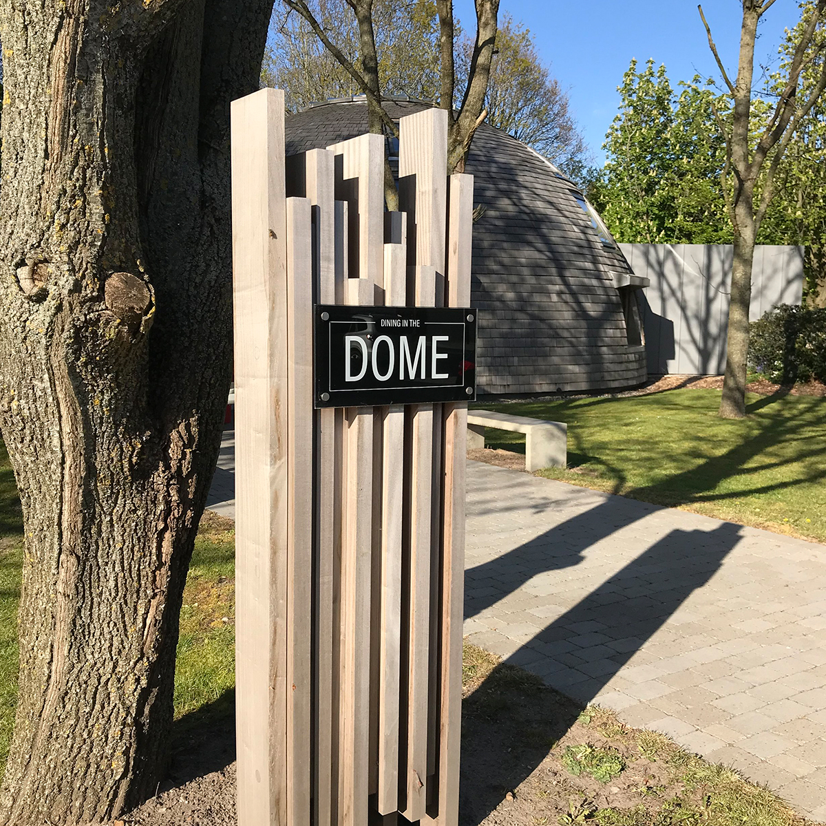 Udendørs wayfinding- eller områdeskilt i træ med teksten ‘DOME’, placeret langs en sti i grønt udemiljø