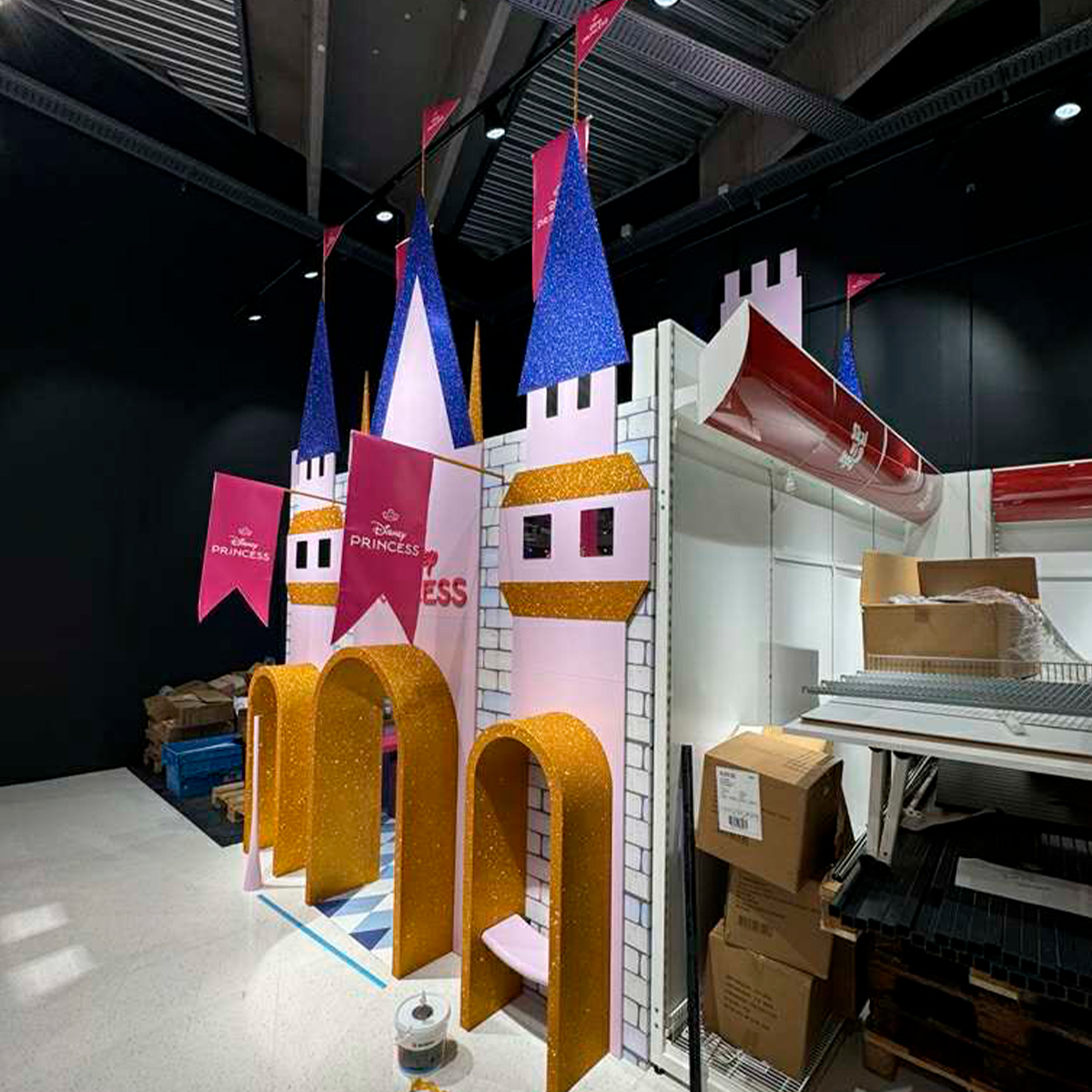 Farverigt specialbygget messe- eller butiksdisplay formet som et eventyrslot med tårne og buer, opstillet i et lager- eller produktionsmiljø