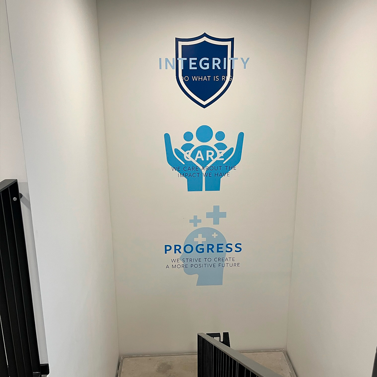 Indendørs væggrafik med logoer og værdierne ‘Integrity’, ‘Care’ og ‘Progress’, placeret i trappeopgang eller gangareal