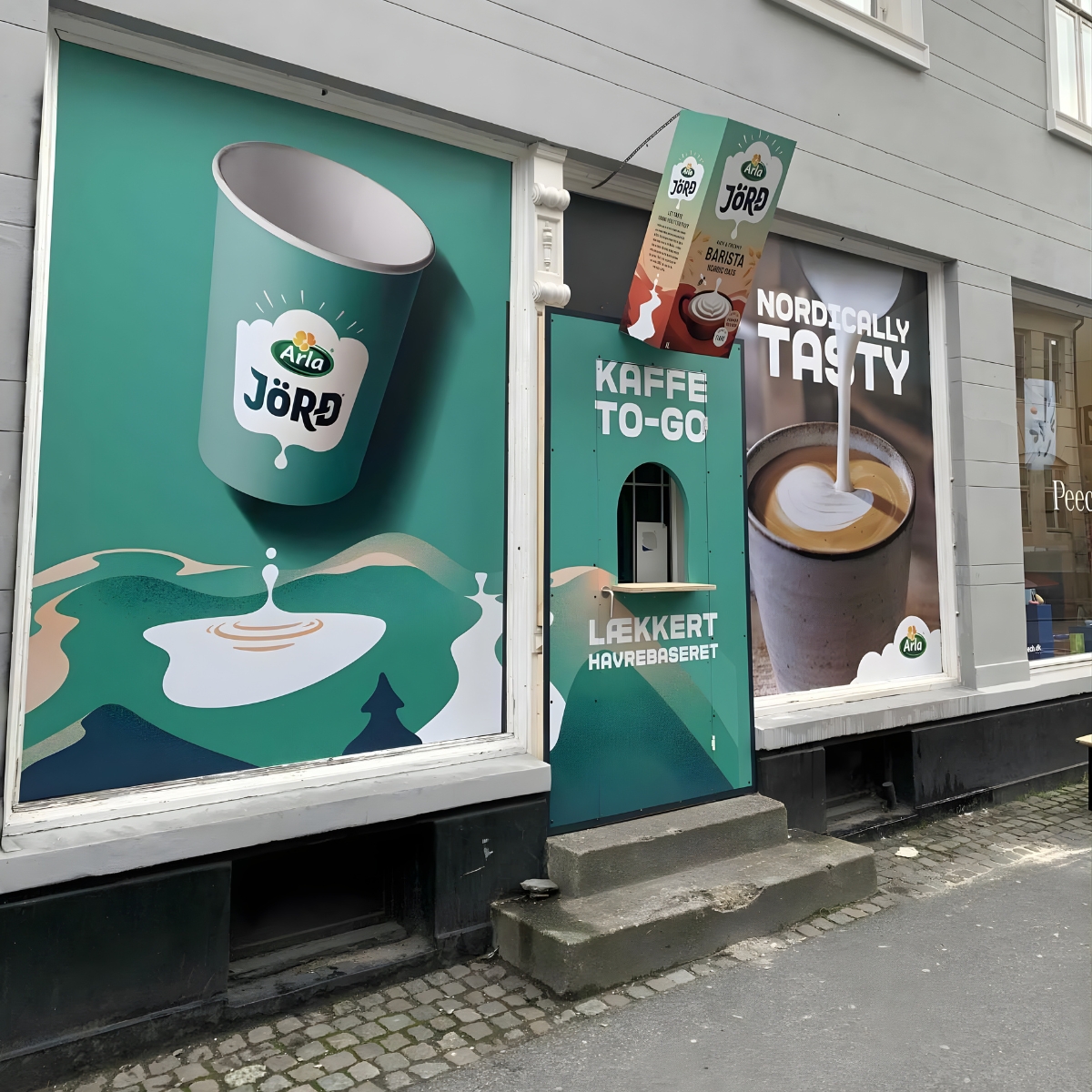 Butiksvinduer med store reklameplakater for kaffe og takeaway, monteret som vinduesgrafik på facade