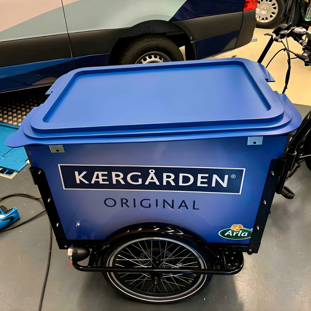 Blå transport- og salgscontainer med teksten KÆRGÅRDEN, monteret på ladcykel i et værksted