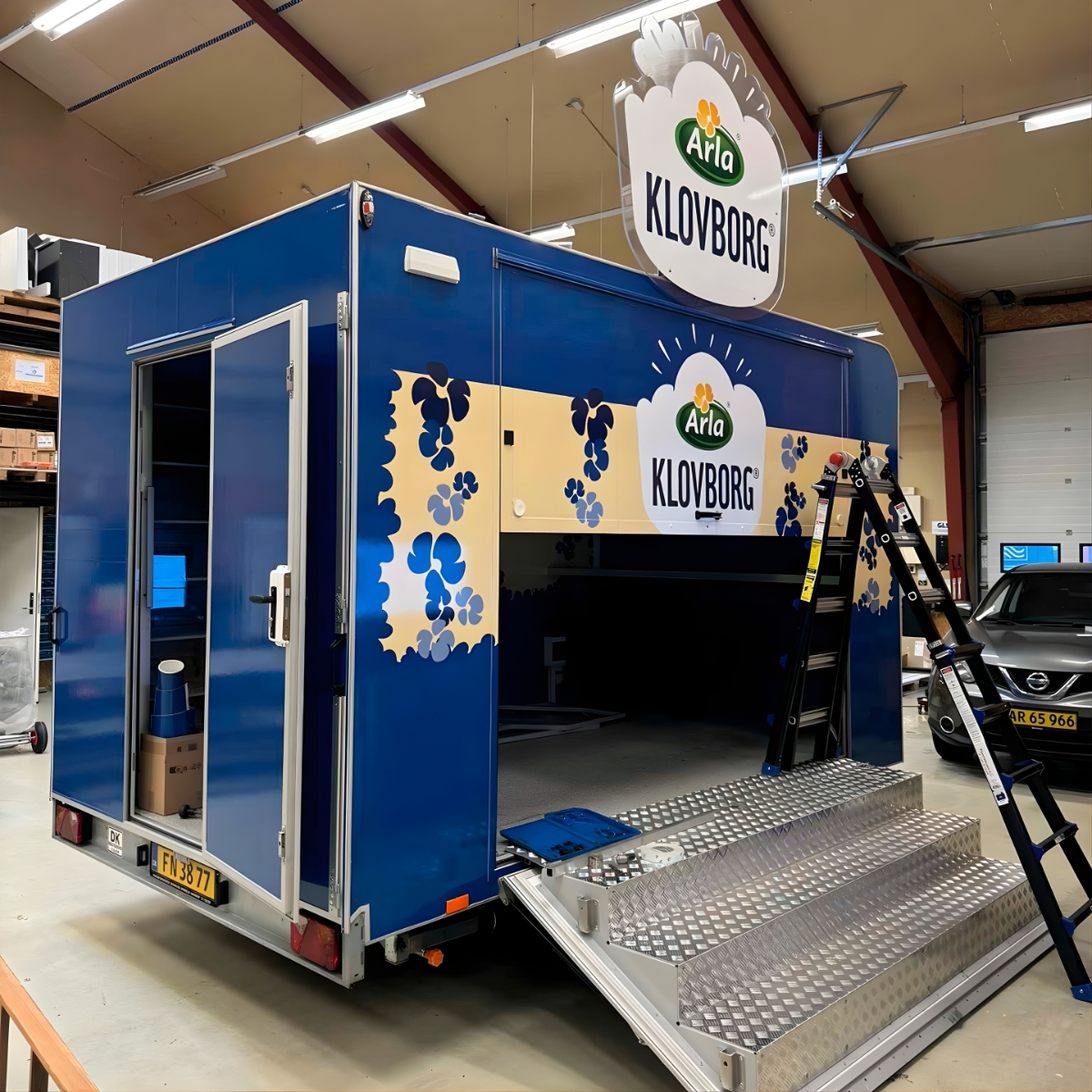 Blå specialbygget salgs- og eventtrailer med Klovborg ost branding, rampe og serveringsåbning, placeret indendørs i produktionsmiljø