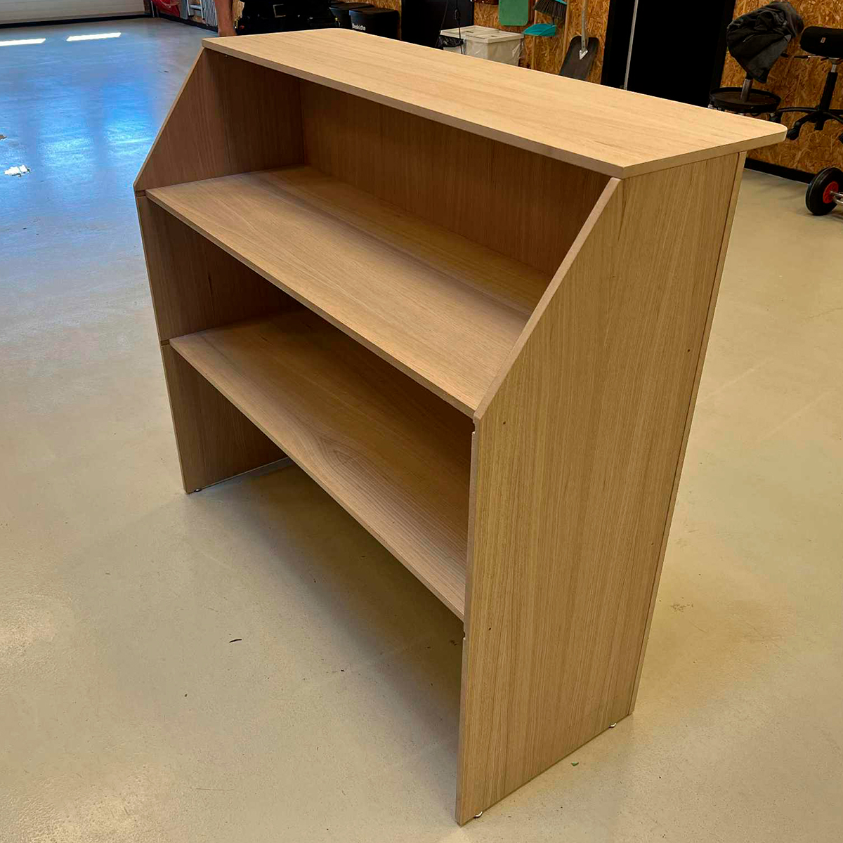 Lille reol i lyst træ med to hylder, placeret fritstående i et værksteds- eller produktionsmiljø