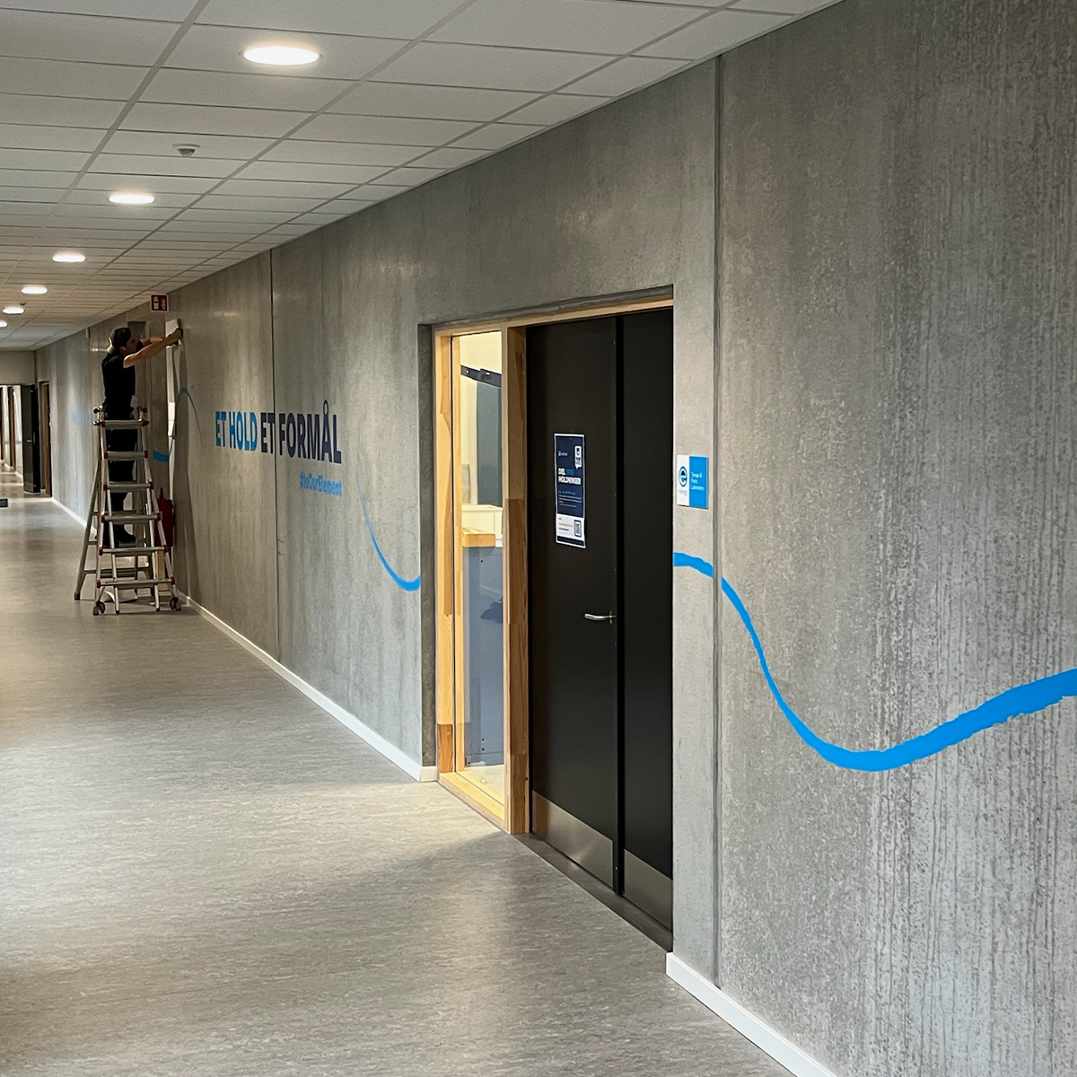 Lang gang med betonvæg med blå wayfinding på væggen 